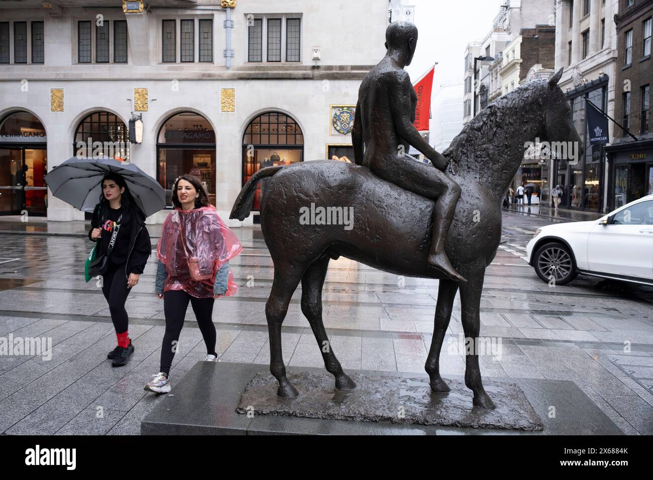 Die Menschen im Regen passieren die Horse and Rider Skulptur von Elizabeth Frink in der Bond Street am 6. Mai 2024 in London, Großbritannien. Bond Street ist eine der wichtigsten Straßen im West End Einkaufsviertel und ist sehr hochwertig. Es ist seit dem 18. Jahrhundert eine modische Einkaufsstraße. Das reiche und wohlhabende Geschäft hier ist hauptsächlich für High-End Mode und Schmuck. Stockfoto