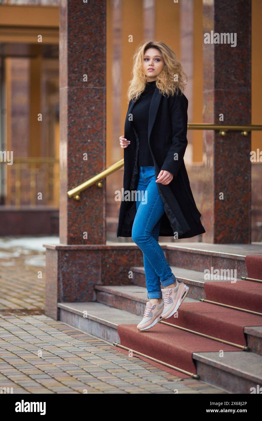 Porträt eines schönen jungen, schlanken, kaukasischen lockigen blonden Mädchens in schwarzem Trenchcoat und blauer Jeans, der die Treppe mit etwas Schnee hinuntergeht Stockfoto