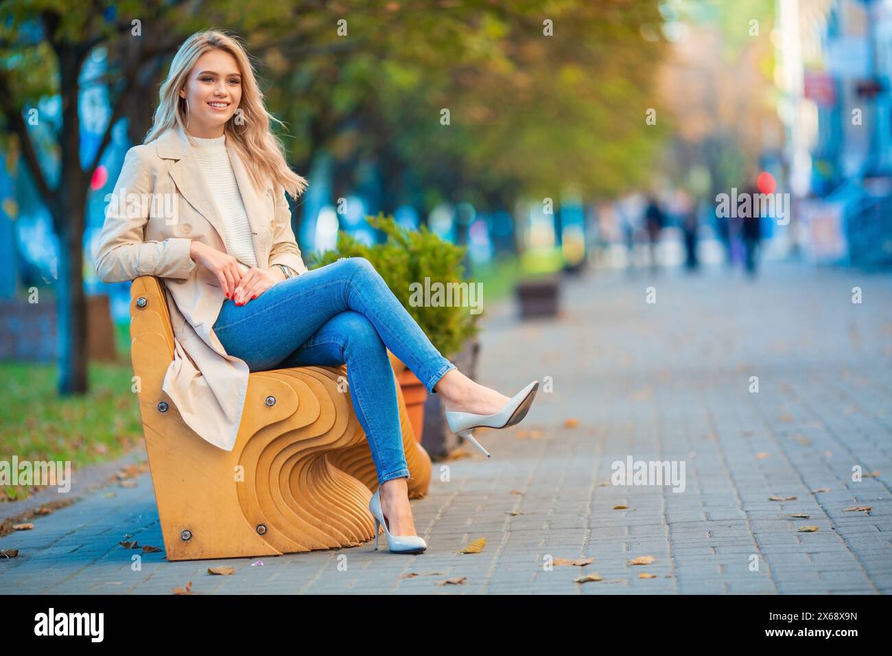 Horizontales Porträt eines lächelnden jungen blonden Mädchens, das entspannt auf einer Holzbank sitzt und blaue Jeans und einen beigen Trenchcoat trägt Stockfoto