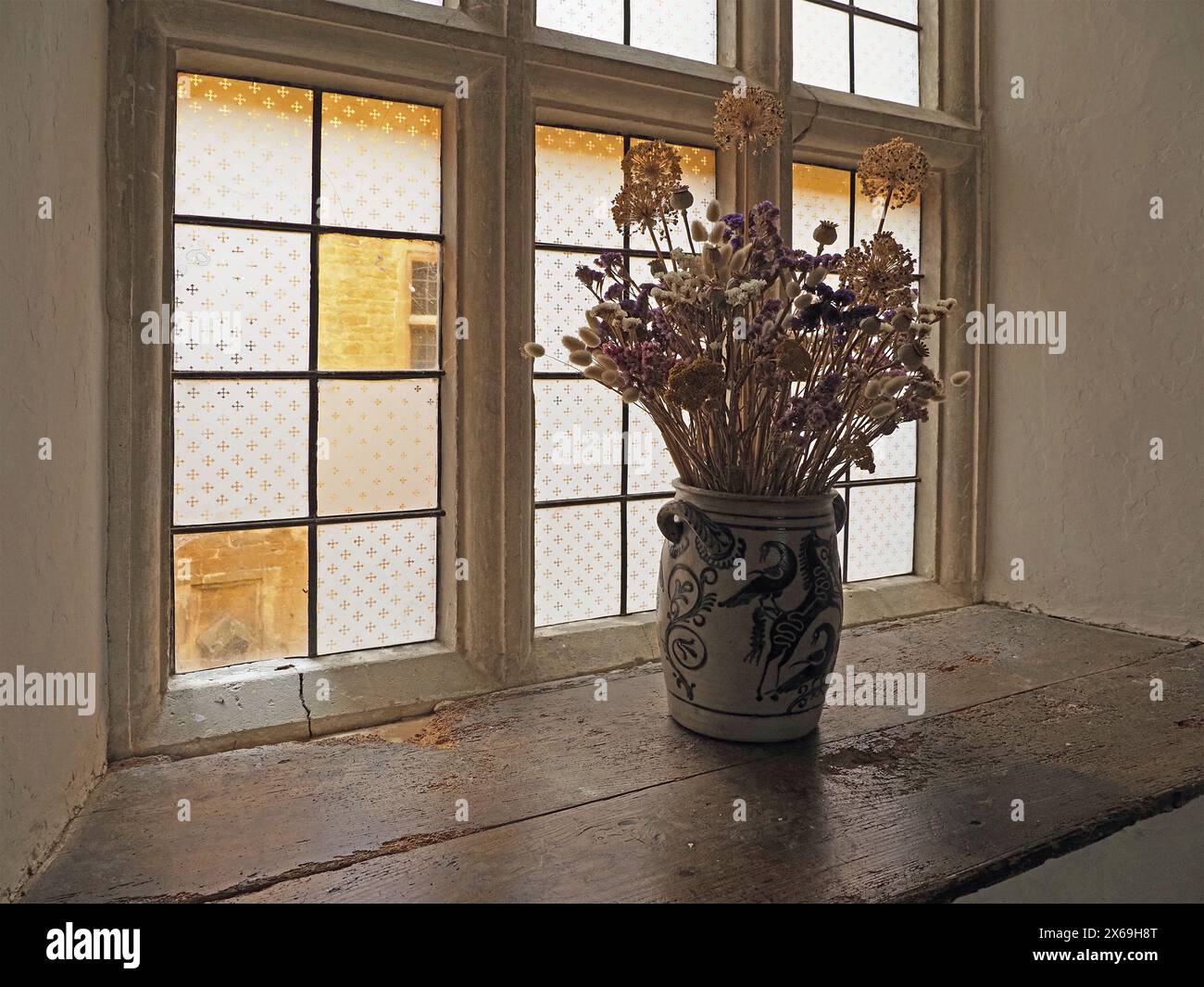 Sonnenlicht strömt durch farbig gemustertes Glas des antiken Fensters in einem historischen Haus mit verfallenem Holz in Fensterbank und Vase mit getrockneten Blumen Stockfoto