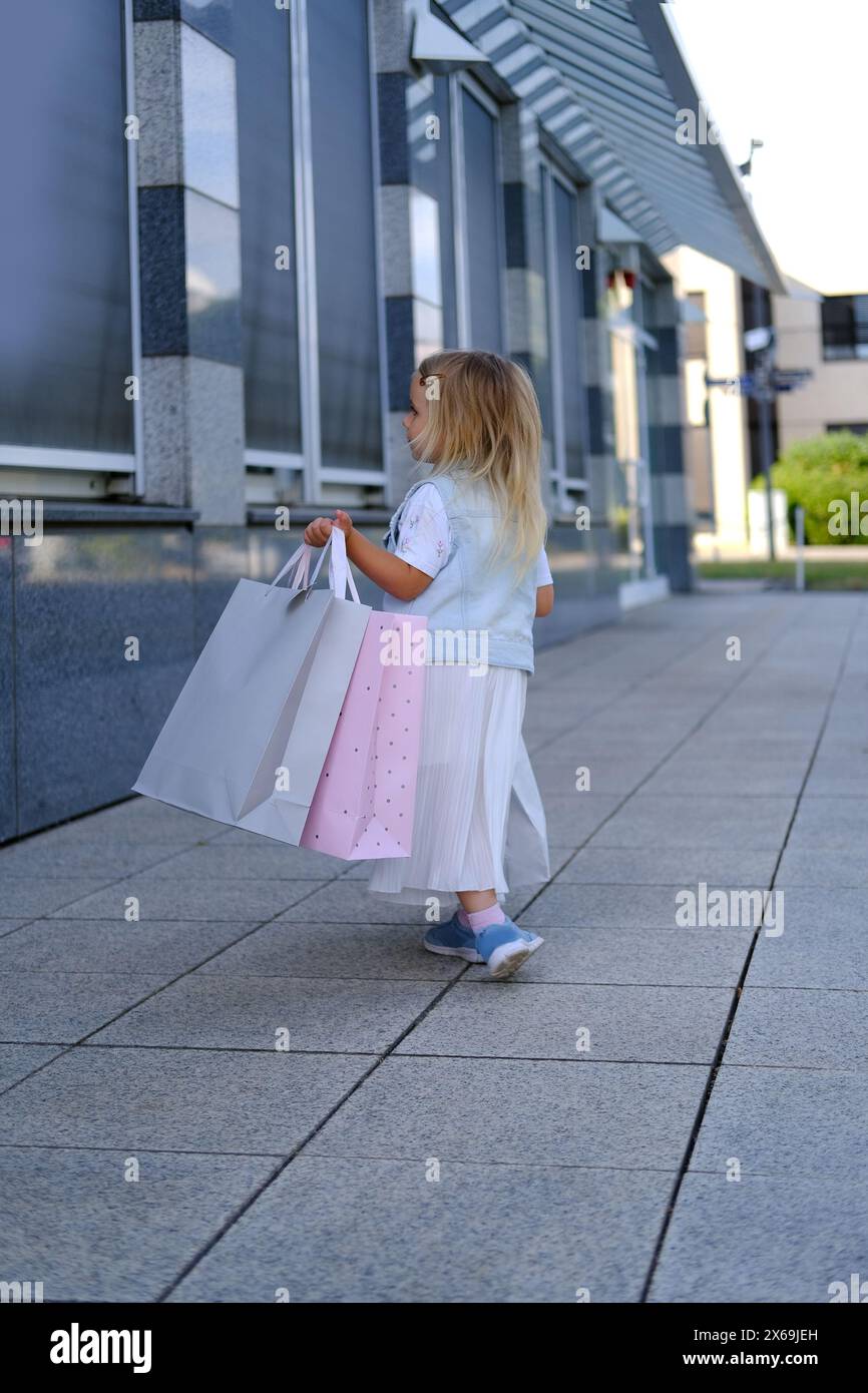 Glückliches Kind von 3-4 Jahren geht aus dem Laden, Mädchen in leichten modischen Kleidern, Bastelpapiertüten voller Einkäufe in beiden Händen, glückliches Einkaufen auf B Stockfoto