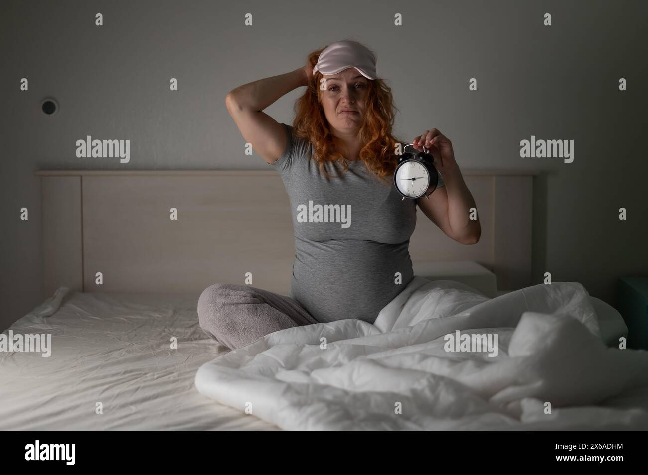Eine Kaukasische Schwangere sitzt im Bett und hält einen Wecker. Schlaflosigkeit. Stockfoto