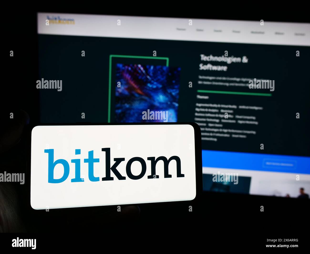 Person mit Smartphone mit Logo des deutschen Telekommunikationsverbandes Bitkom e. V. vor der Website. Schwerpunkt auf dem Display des Telefons. Stockfoto