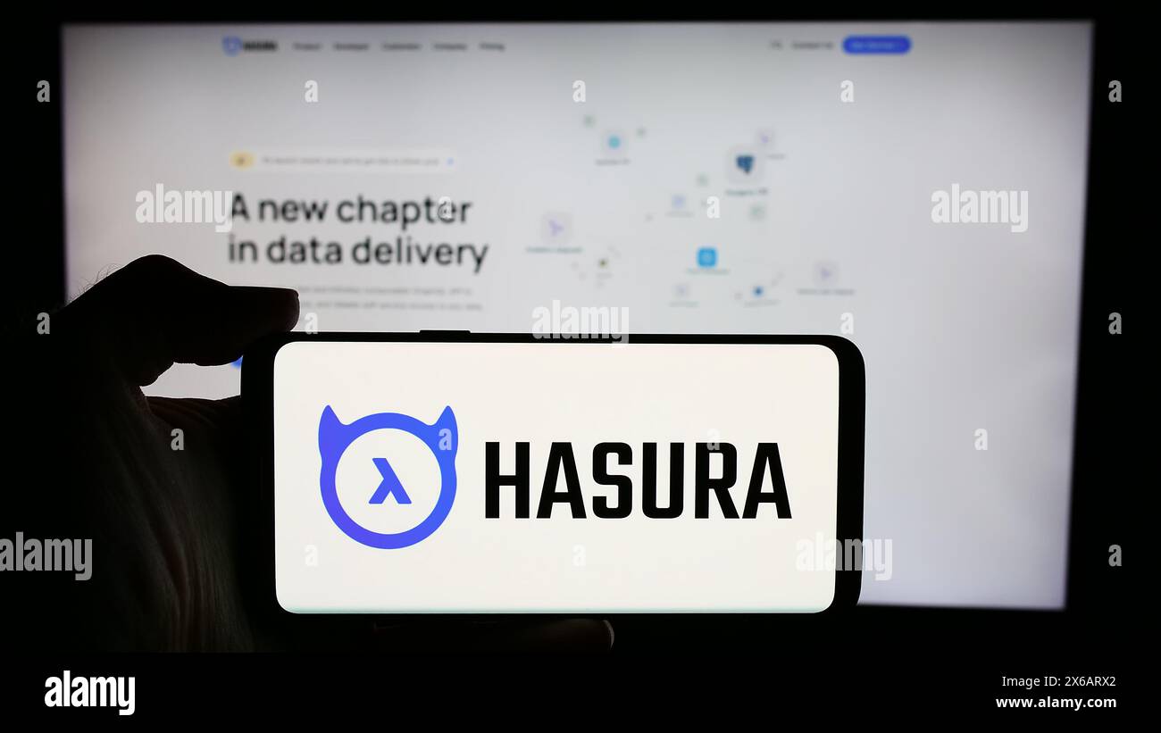 Person, die ein Mobiltelefon mit dem Logo des amerikanischen Softwareentwicklungsunternehmens Hasura Inc. Vor der Business-Webseite hält. Schwerpunkt auf dem Display des Telefons. Stockfoto