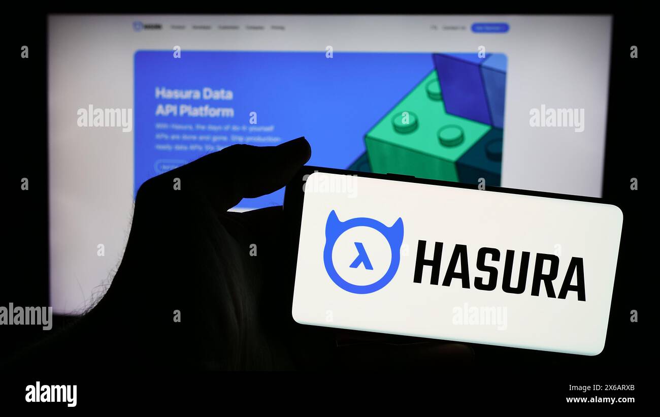 Person, die ein Mobiltelefon mit dem Logo des US-Softwareentwicklungsunternehmens Hasura Inc. Vor der Website des Unternehmens hält. Schwerpunkt auf dem Display des Telefons. Stockfoto