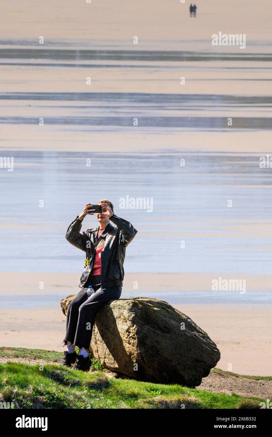 Eine Frau, die allein auf einem Felsen sitzt und ein Smartphone benutzt, um ein Selfie bei Fistral in Newquay in Cornwall in England zu machen. Stockfoto