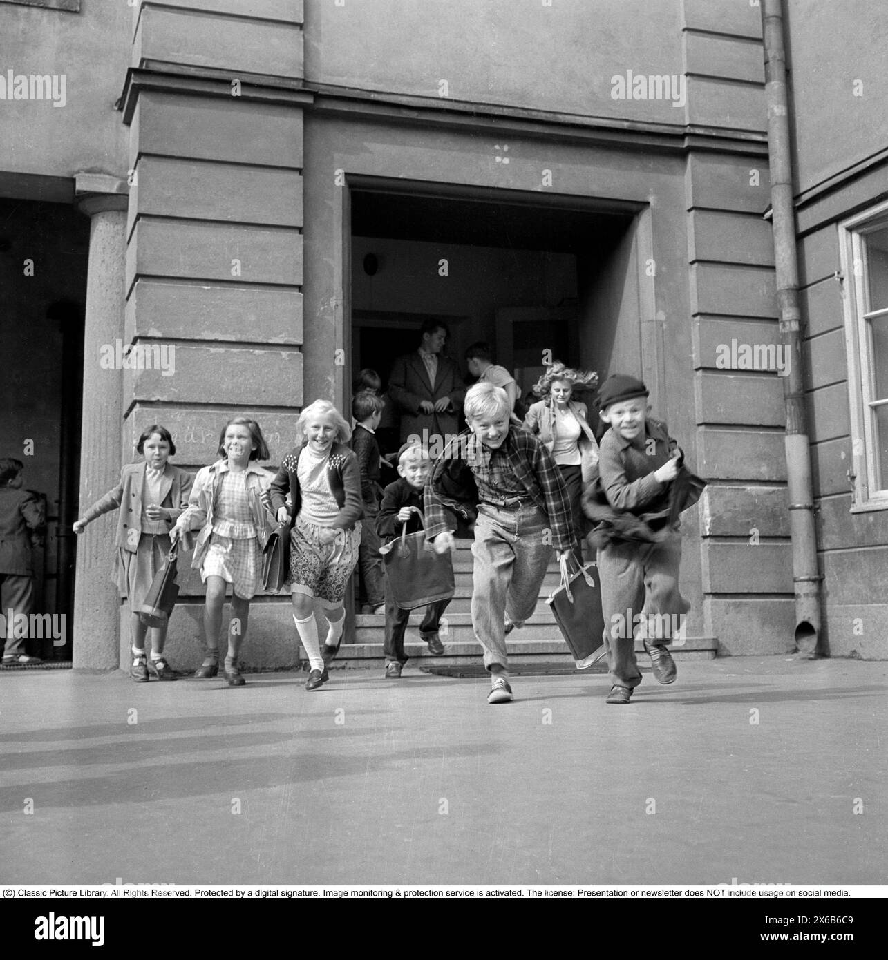 Schulkinder der 1950er Jahre. Der Schultag oder gar die Schulzeit könnte zu Ende sein und die Kinder laufen glücklich aus dem Schulgebäude. Roland Palm Ref. 10-82-1 Stockfoto