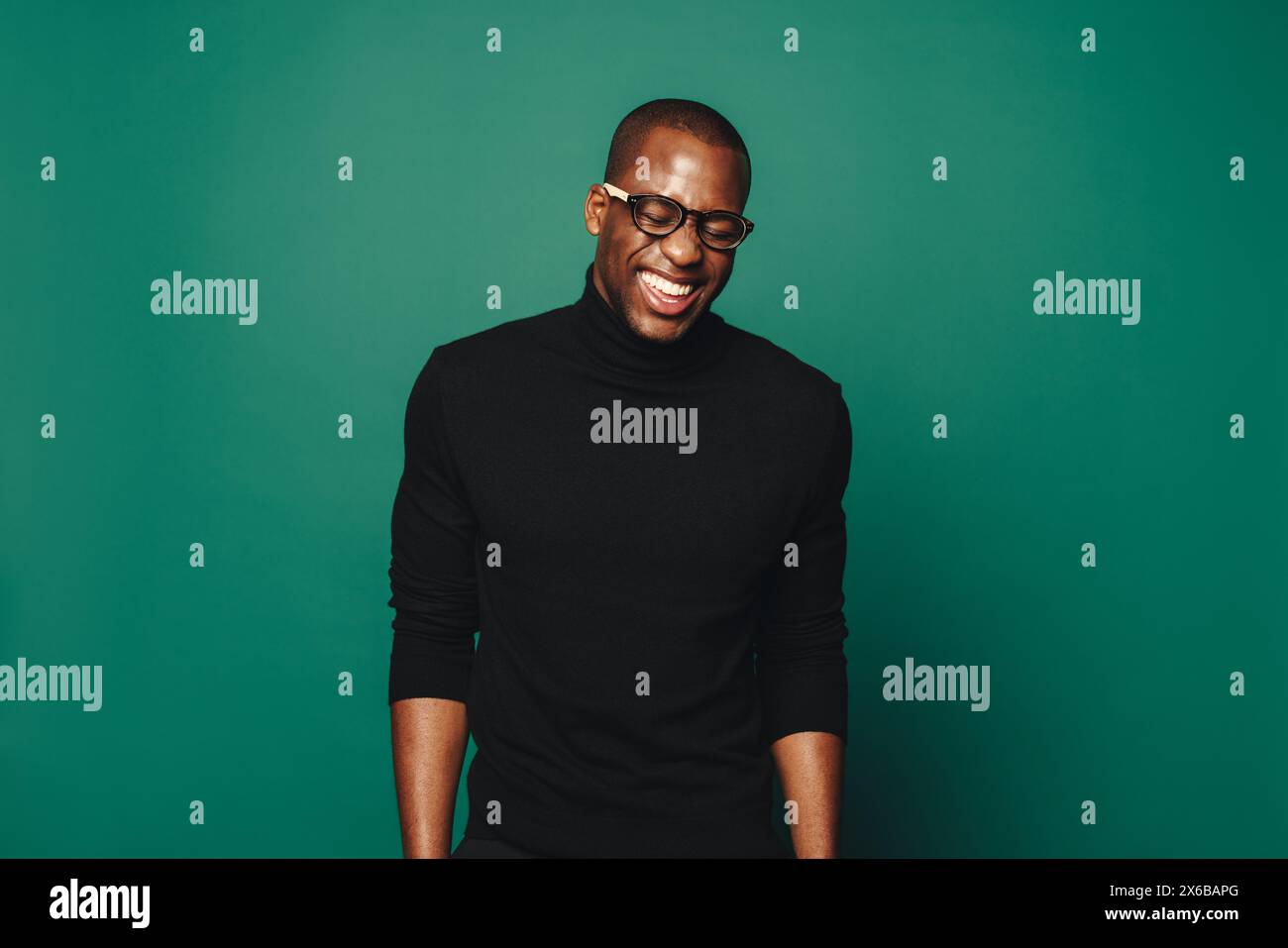 Ein junger afrikanischer Mann mit einem selbstbewussten Lächeln steht in einem Studio. Er trägt eine Brille und einen lässigen schwarzen Pullover, der auf dem grünen Rücken Glück ausstrahlt Stockfoto