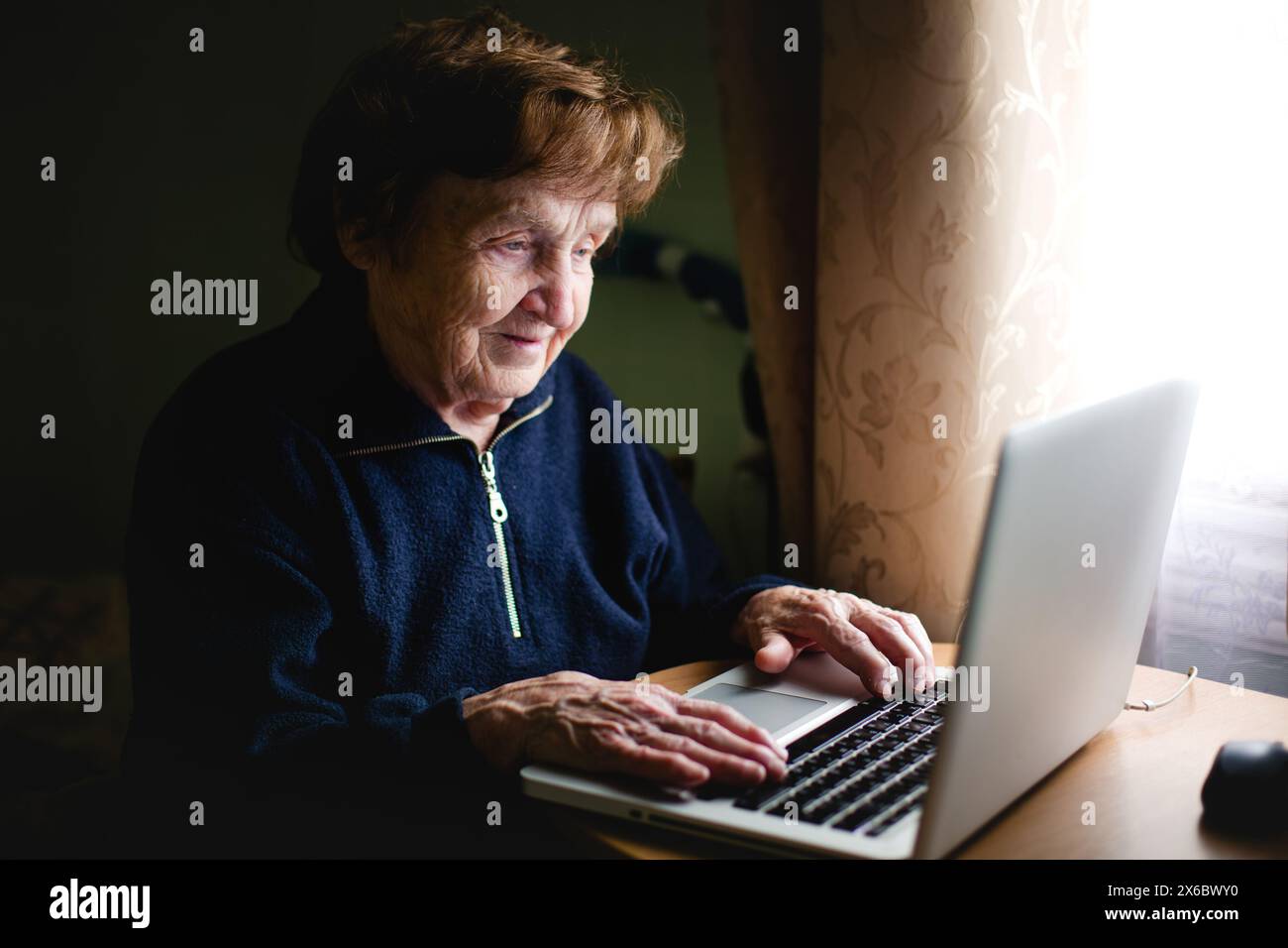 Eine ältere Frau sitzt mit einem Laptop an einem Schreibtisch, während sie arbeitet, mit dem Finger über der Tastatur. Stockfoto
