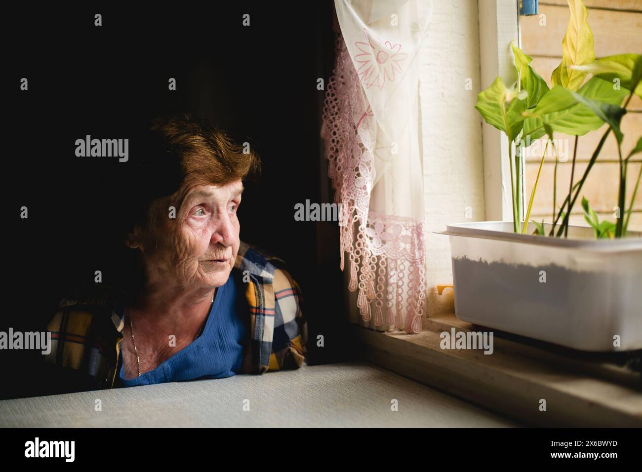 Eine ältere Frau sitzt am Fenster, ihr Blick verloren in der Ferne und erinnert an vergangene Tage. Stockfoto