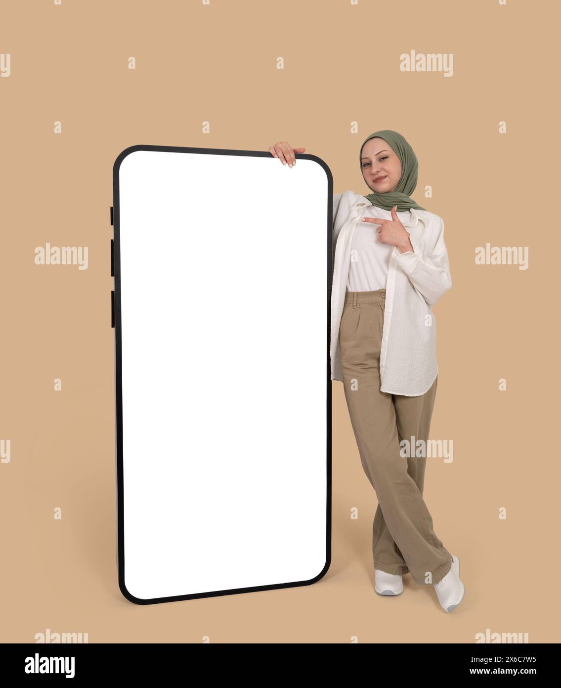 Voller Körper fröhliches kaukasisches muslimisches Mädchen im Hijab, das in der Nähe steht und mit dem Finger auf ein riesiges Handy zeigt. Leerer weißer Bildschirm. Stockfoto