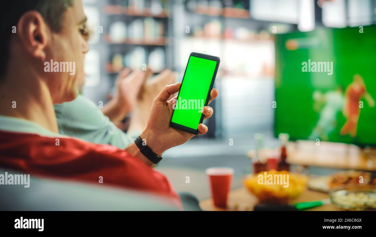 Zu Hause Fußball-Fan hält Green Screen Chroma Key Smartphone, Freunde sehen Fußball-Spiel im Fernsehen, feuern Favourtite Sports Team an, um die Meisterschaft zu gewinnen. Football Club Play. Über der Schulter Stockfoto