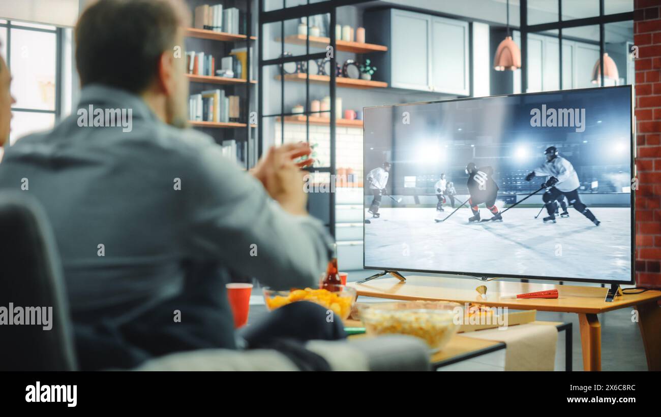 Zu Hause sitzen Eishockeyfans auf einer Couch und schauen sich das Spiel im Fernsehen an, wenn das Favourtite Sports Team die Meisterschaft gewinnt. Der Bildschirm Zeigt Professionelle Spieler Während Der Weltmeisterschaft. Über der Schulter Stockfoto