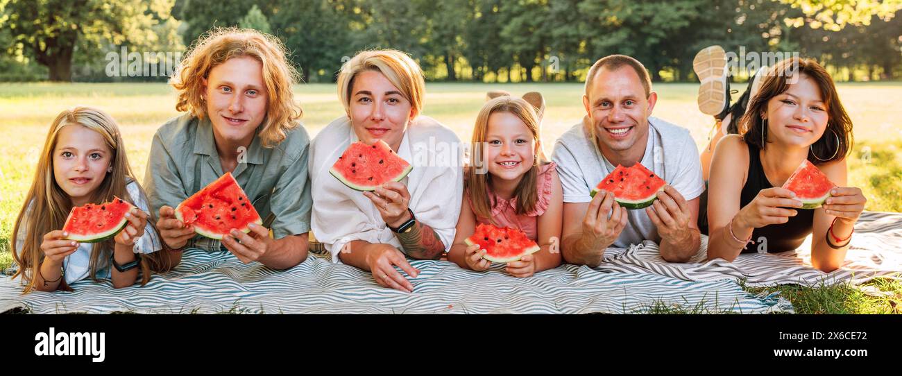 Große Familie, die auf der Picknickdecke im Stadtpark unter Linden Tree am Wochenende liegt, sonntags am Sonnentag, lächelt, vor der Kamera lacht und Wasser isst Stockfoto