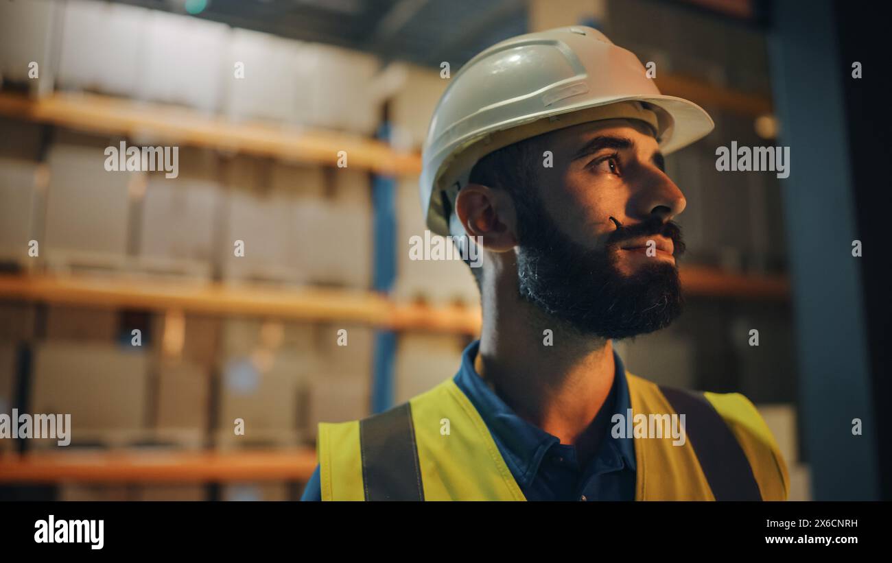 Porträt eines männlichen lateinischen Arbeiters, der einen harten Hut trägt und vor dem Lager lächelnd steht. Professioneller Frontline-Held mit stilvollem Bart im Kinolicht Stockfoto