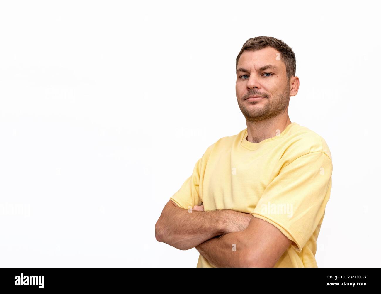 Isoliertes Porträt eines Mannes, der ein gelbes T-Shirt trägt, mit überkreuzten Armen steht, lächelt und in die Kamera blickt. Isoliert auf weißem Hintergrund. Stockfoto