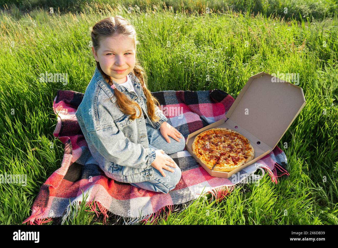 Ein Mädchen in einem blauen Denim-Anzug mit einer Pizzaschachtel sitzt auf einer Decke auf dem grünen Gras. Sonniger Sommertag. Picknick im Freien Stockfoto