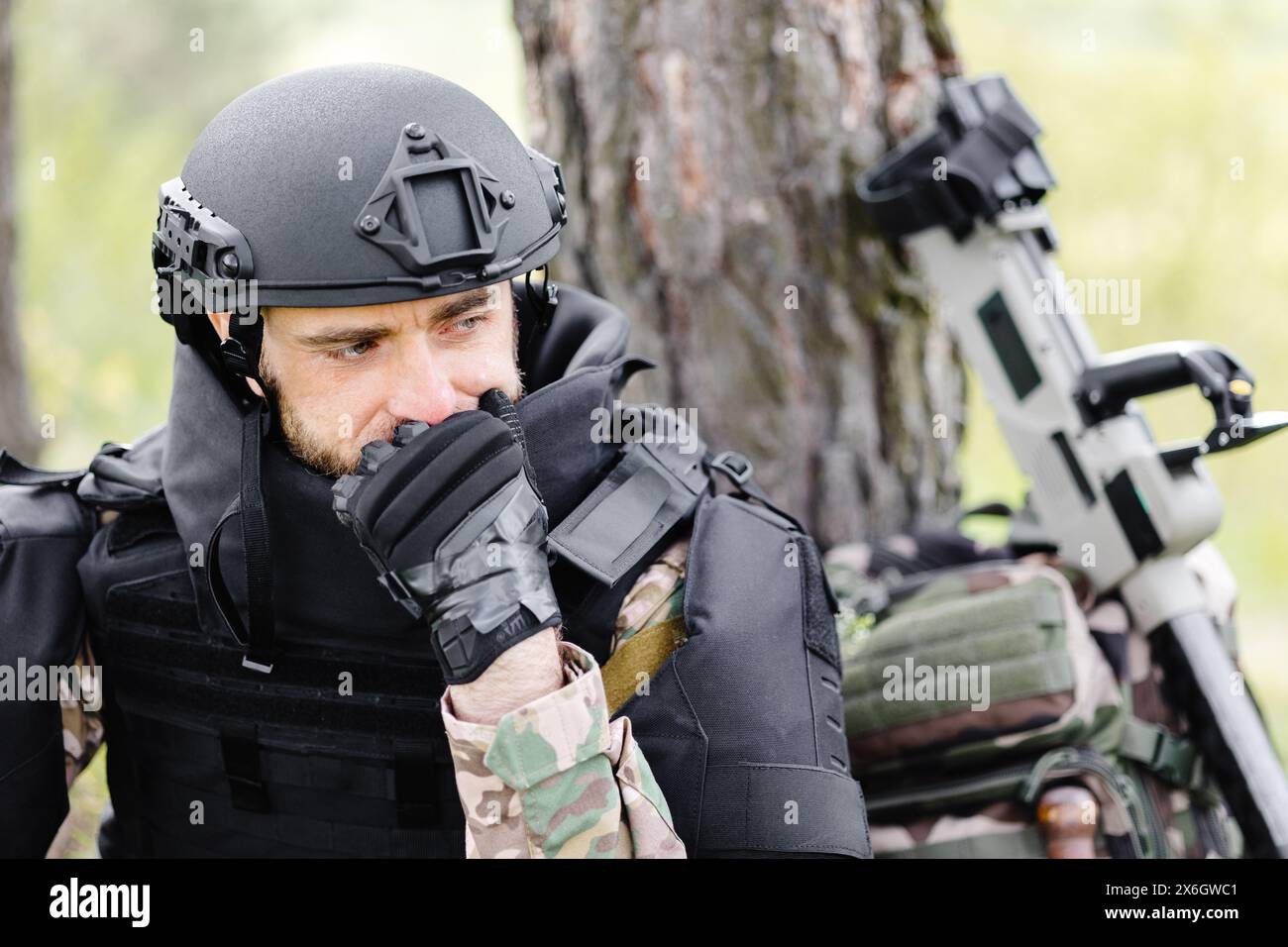 Ein Mann in Militäruniform und kugelsicherer Weste sitzt im Wald neben einem Metalldetektor und einem Militärrucksack. Ein Mann hält an der Arbeit des Dämons Stockfoto