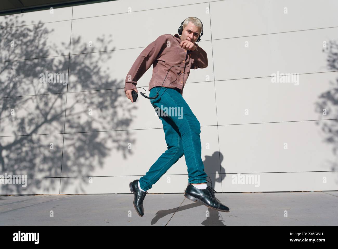 Junger lateinamerikanischer Mann mit kurzen blonden Haaren in Retro-Kleidung, Hosen und braunem Hemd, draußen springt er mit Kopfhörern Stockfoto