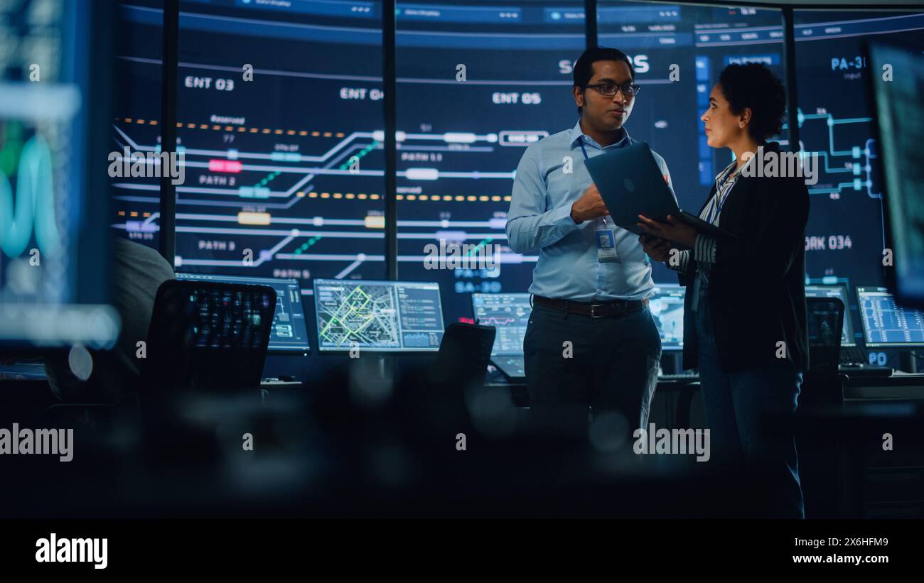 Im System Control Room führen Projektmanager und IT Technical Engineer mit Laptop Gespräche, umgeben von mehreren Monitoren mit Grafik. Big Monitor Zeigt Informationen Zu Interactive Server Blockchain An. Stockfoto