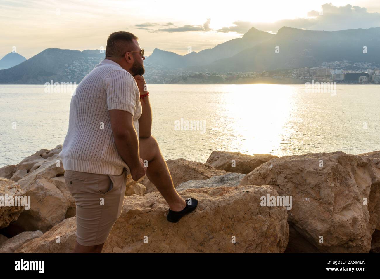 Dieses Porträt fängt einen 30-jährigen Mann ein, während er über die ruhige Weite des Mittelmeers reflektiert. Die untergehende Sonne wirft ein goldenes Leuchten über den Stockfoto