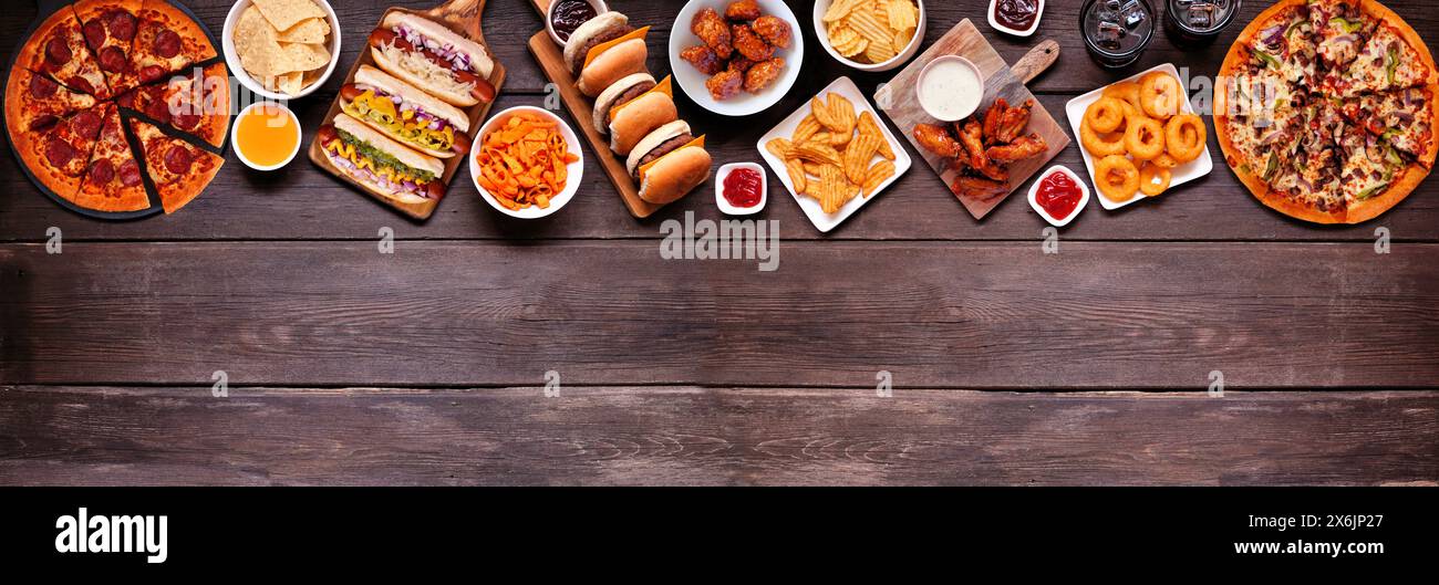 Junk Food obere Grenze. Pizza, Hot Dogs, Hamburger, Hähnchenflügel und salzige Snacks. Über dem Hintergrund eines dunklen Holzbanners mit Kopierraum. Stockfoto