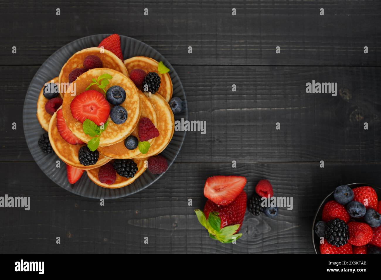 Ein flacher goldener Pfannkuchen auf schwarzem Holzhintergrund mit frischen Beeren sorgt für eine gemütliche und verwöhnende Szene. Stockfoto