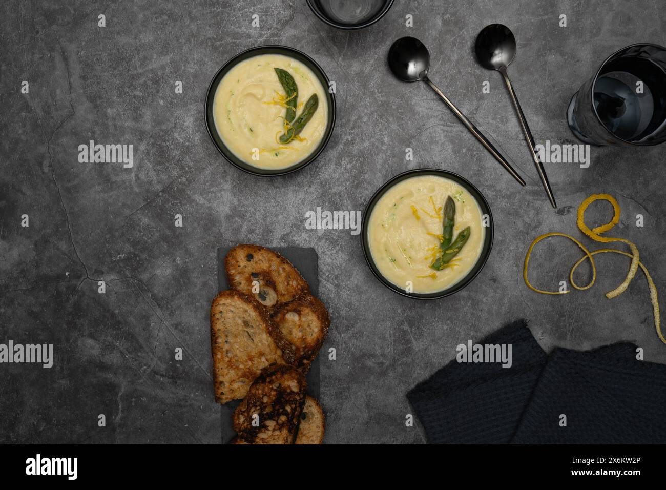 Auf einem grauen Hintergrund werden Schüsseln mit cremiger spargelsuppe, geschmückt von grünem spargel, von Brottoasts, eleganten Löffeln flankiert Stockfoto