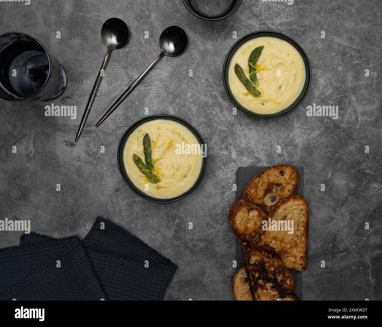 Auf einem grauen Hintergrund werden Schüsseln mit cremiger spargelsuppe, geschmückt von grünem spargel, von Brottoasts, eleganten Löffeln flankiert Stockfoto