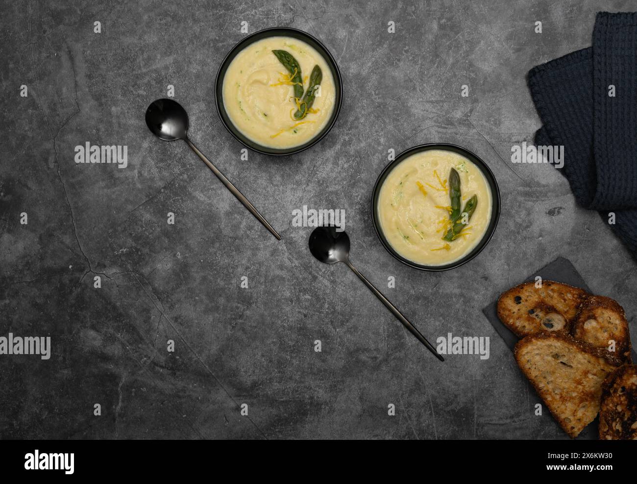 Auf einem grauen Hintergrund werden Schüsseln mit cremiger spargelsuppe, geschmückt von grünem spargel, von Brottoasts, eleganten Löffeln flankiert Stockfoto