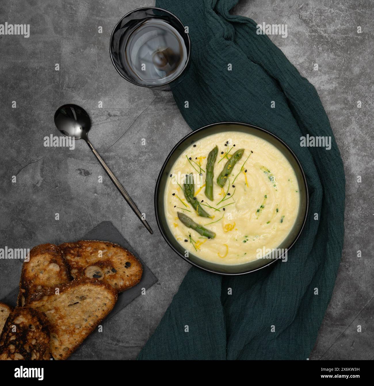 Cremeweiße Spargelsuppe auf dunkelgrauem Steinhintergrund, umgeben von grünem Küchentuch Stockfoto