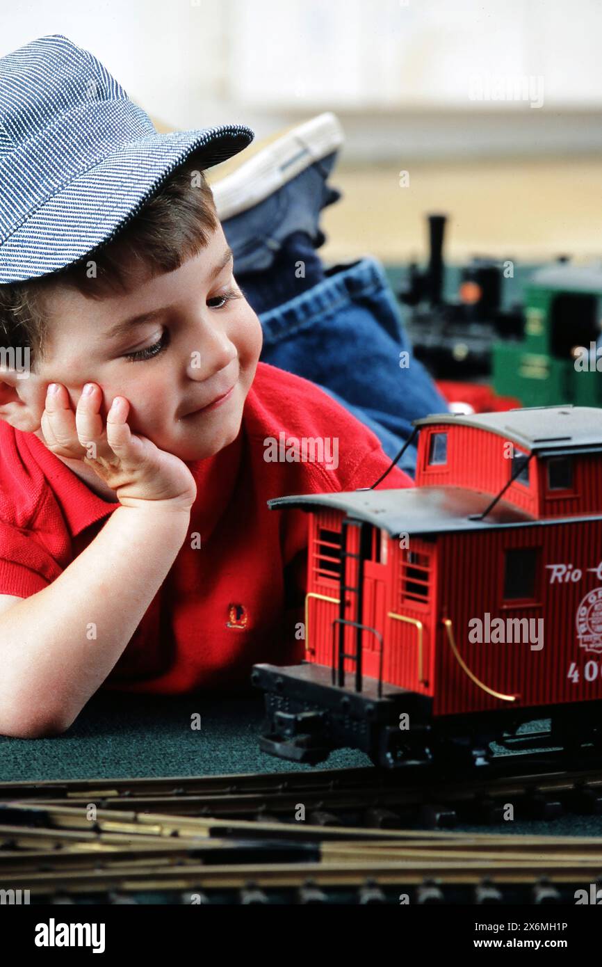 Ein Junge mit einer Ingenieurmütze, der tagträumt, neben einem hellroten Modellzugwagen, der auf dem Boden steht. Der Junge scheint entspannt zu sein und schaut zu Stockfoto