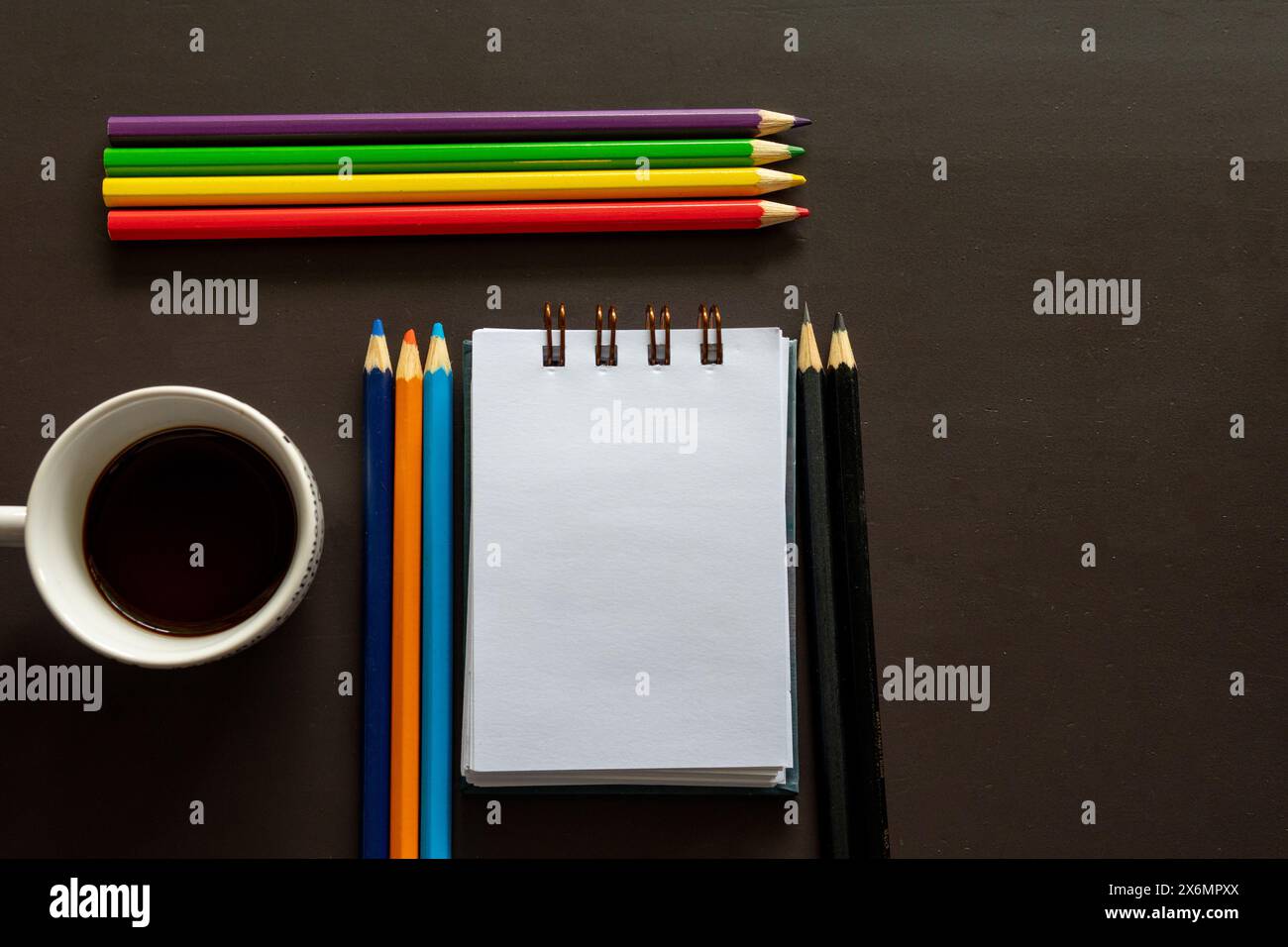 Bunte Bleistifte stehen neben einem Notizblock auf einem dunklen Schreibtisch Stockfoto