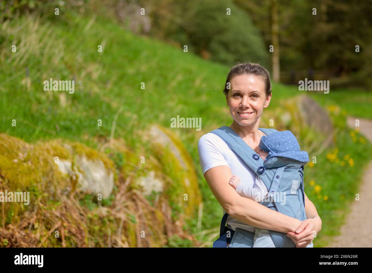 Glückliche Frau, die in die Kamera blickt und lächelt, während sie sie in einer Babytrage im Park hält und trägt Stockfoto