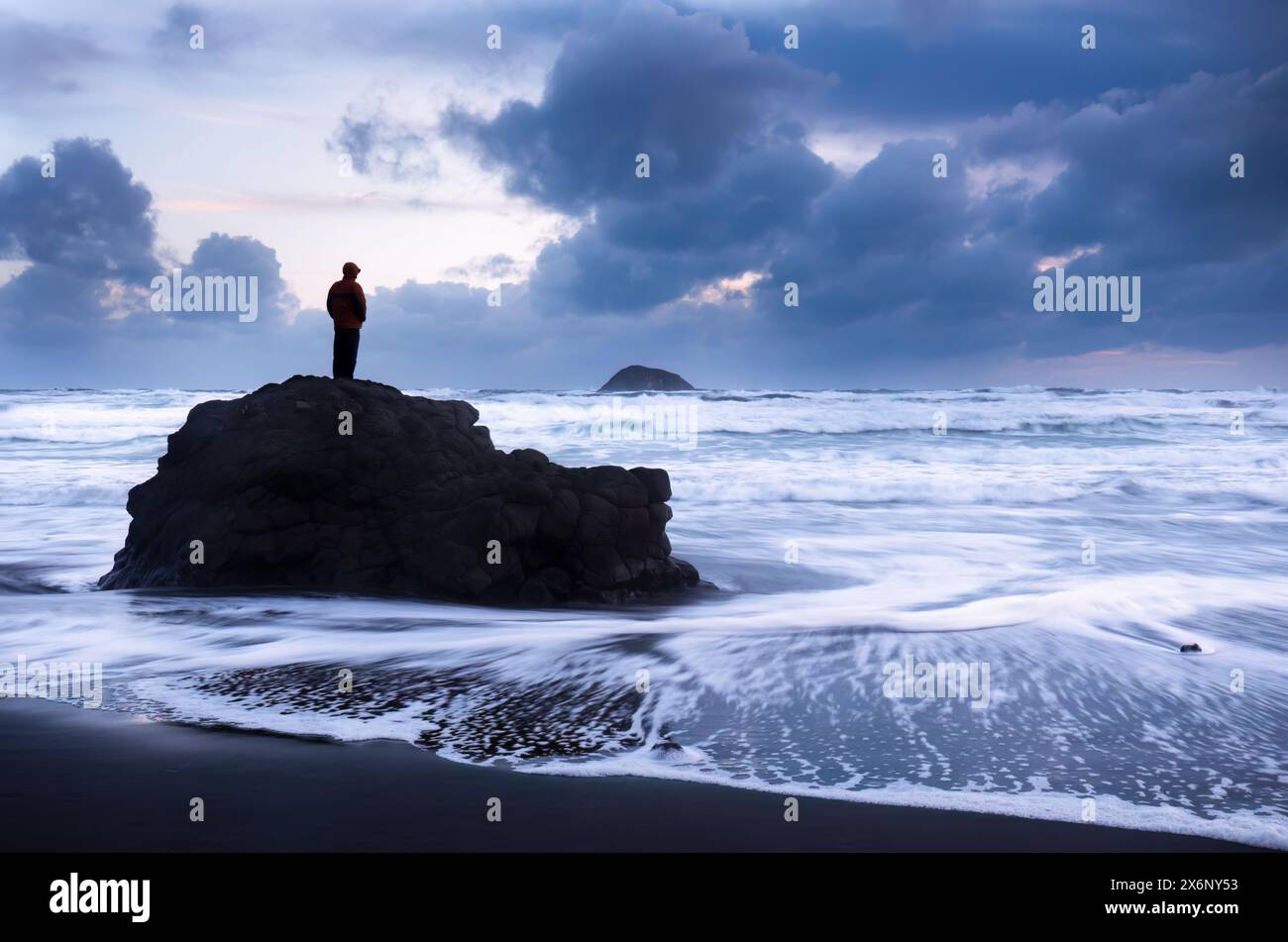 Mann, der auf Felsen steht und auf das stürmische Meer blickt. Oaia Island in der Ferne. Muriwai Beach. Auckland. Stockfoto