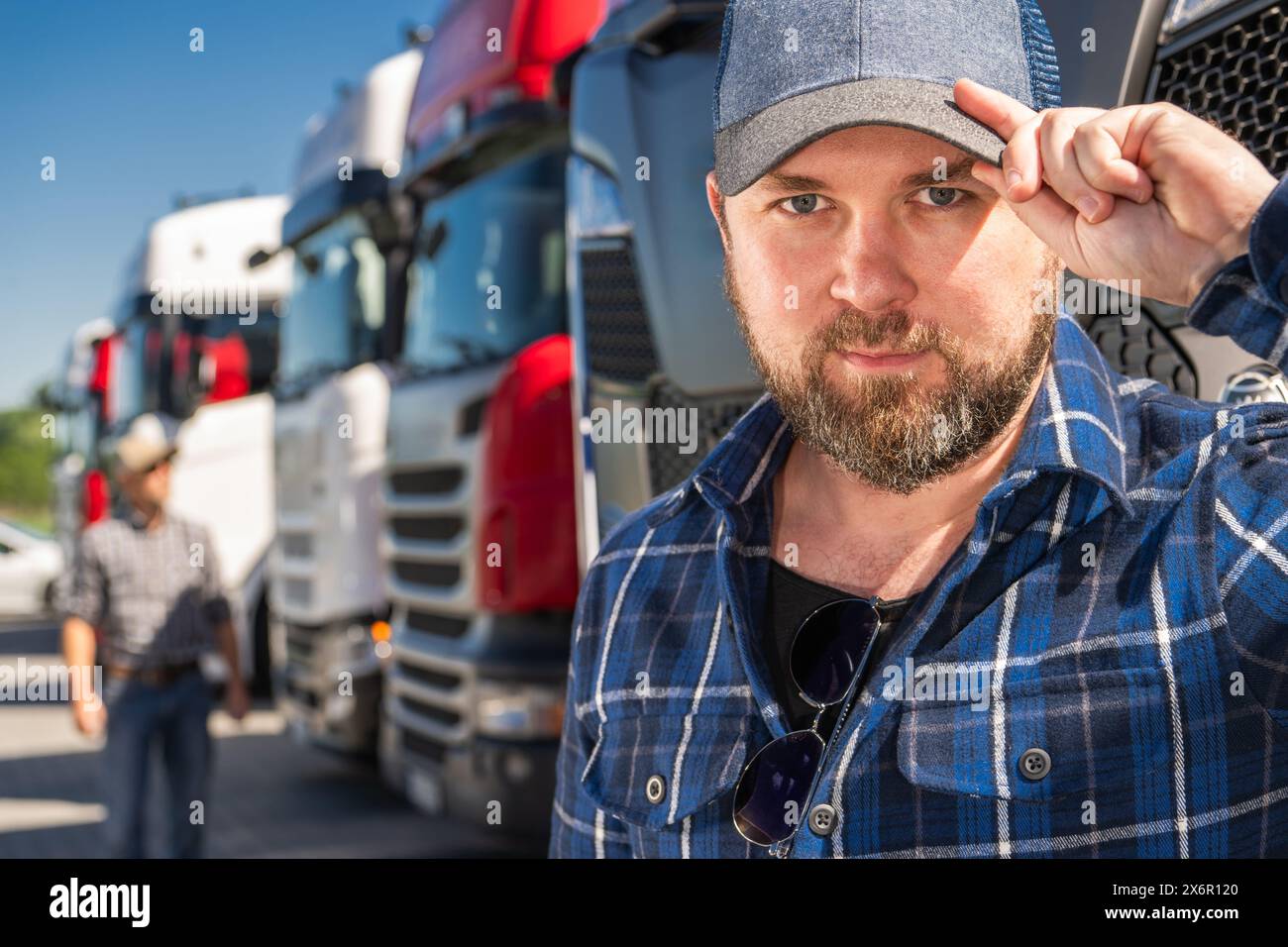 Ein kaukasischer Trucker in seinen 30ern mit Hut steht vor einer Gruppe von Trucks im Euro-Stil. Stockfoto