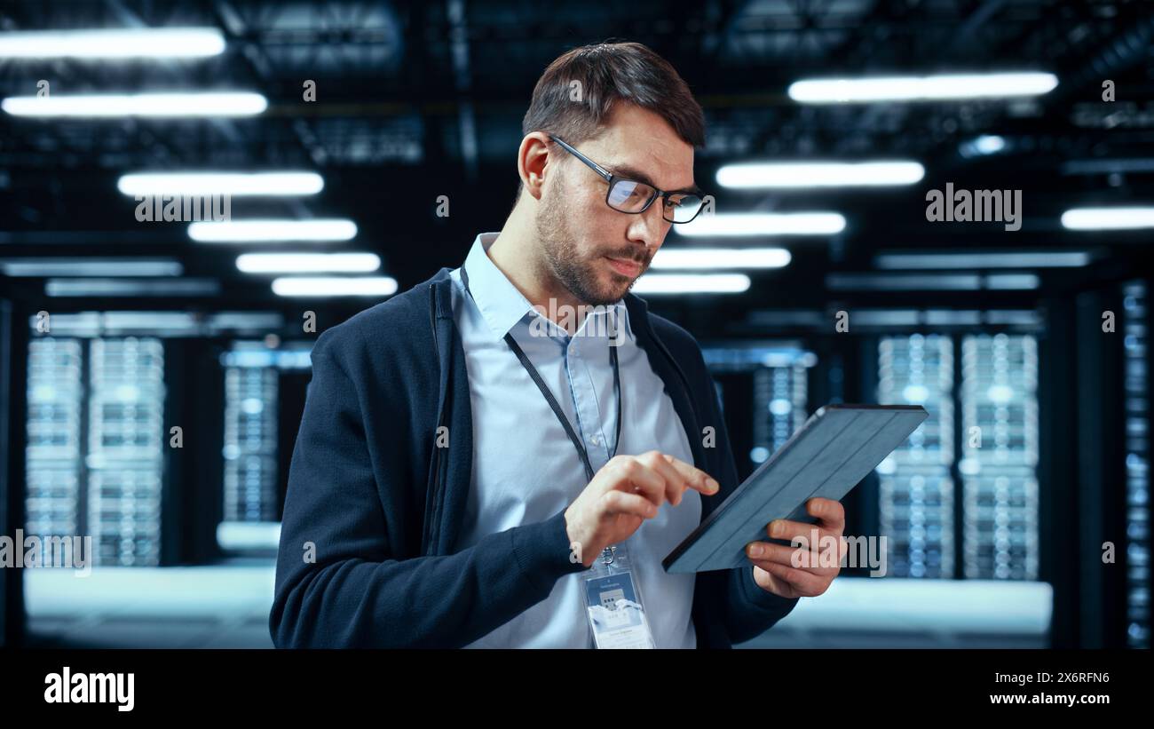 Porträt eines bärtigen, gutaussehenden kaukasischen IT-Ingenieurs in Brille, der mit Tablet steht und in der Mitte eines funktionierenden Serverraums für Rechenzentren posiert, in dem Servercomputer auf einem Rack arbeiten. Stockfoto