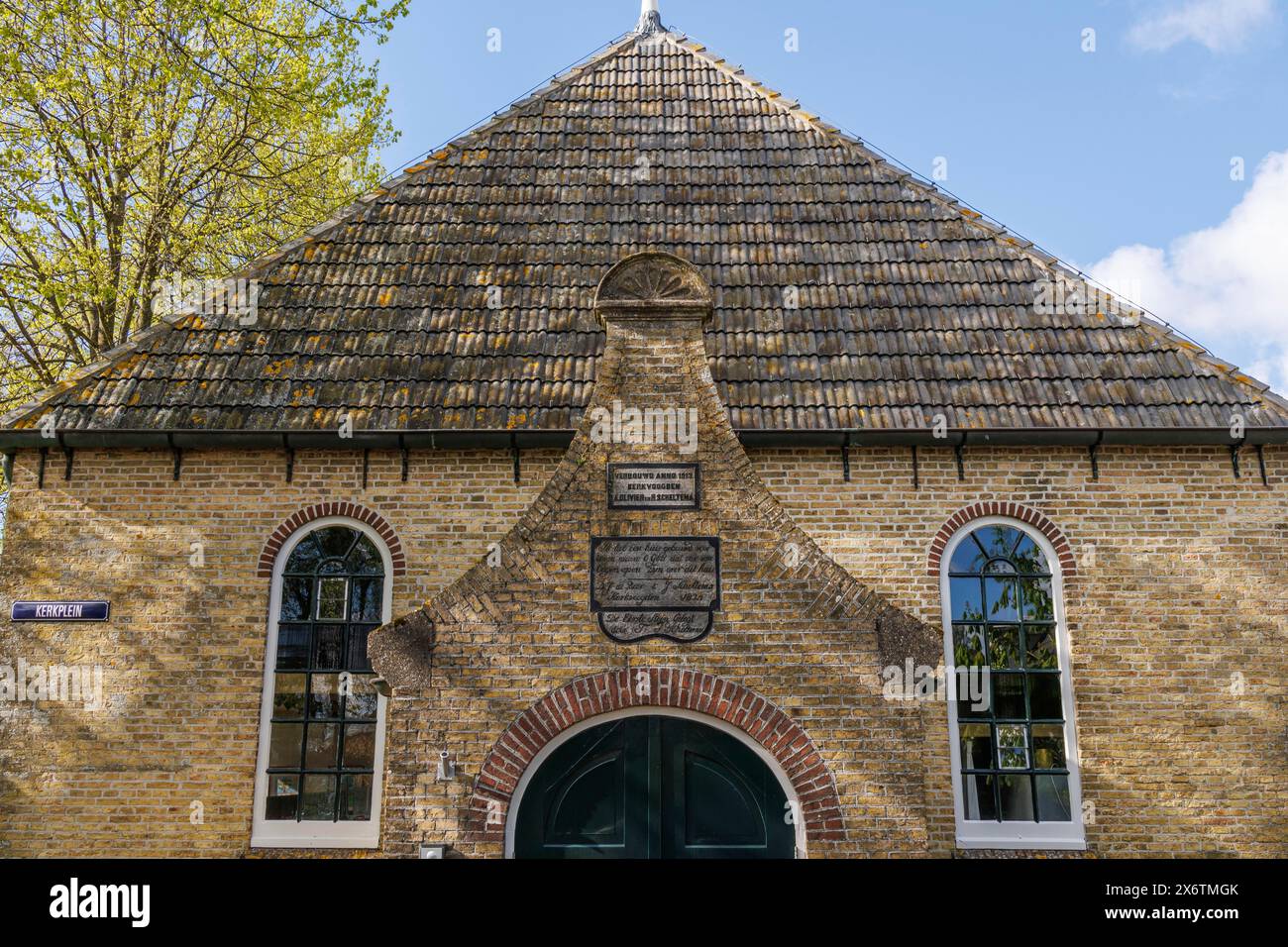 Traditionelles Backsteingebäude mit großem Dach, Fenstern und einem historischen Schild über dem Eingang, alten Häusern und kleinen Straßen mit Laternen und Grün Stockfoto