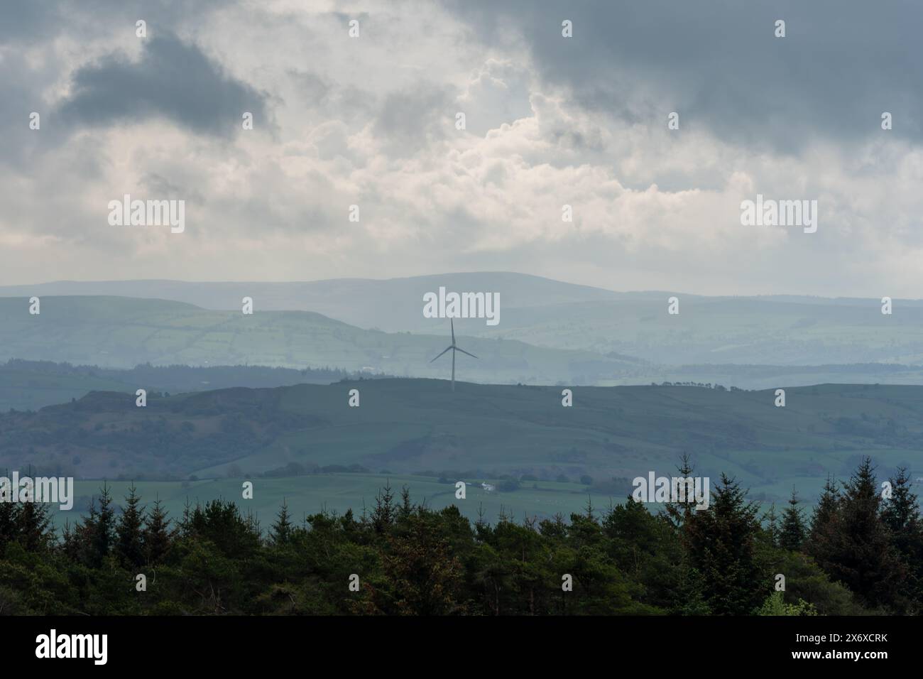 Eine einzelne Windkraftanlage steht auf einem nebelbedeckten Hügel mit grünen Schichten unter einem bewölkten Himmel Stockfoto