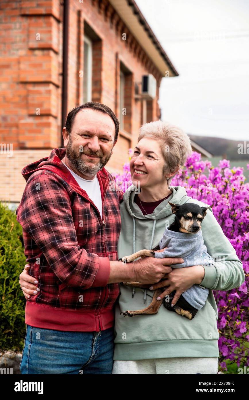 Reifes russisches Ehepaar umarmt sich in der Nähe des Blütenstrauchs von Rhododendron und hält einen Hund Stockfoto