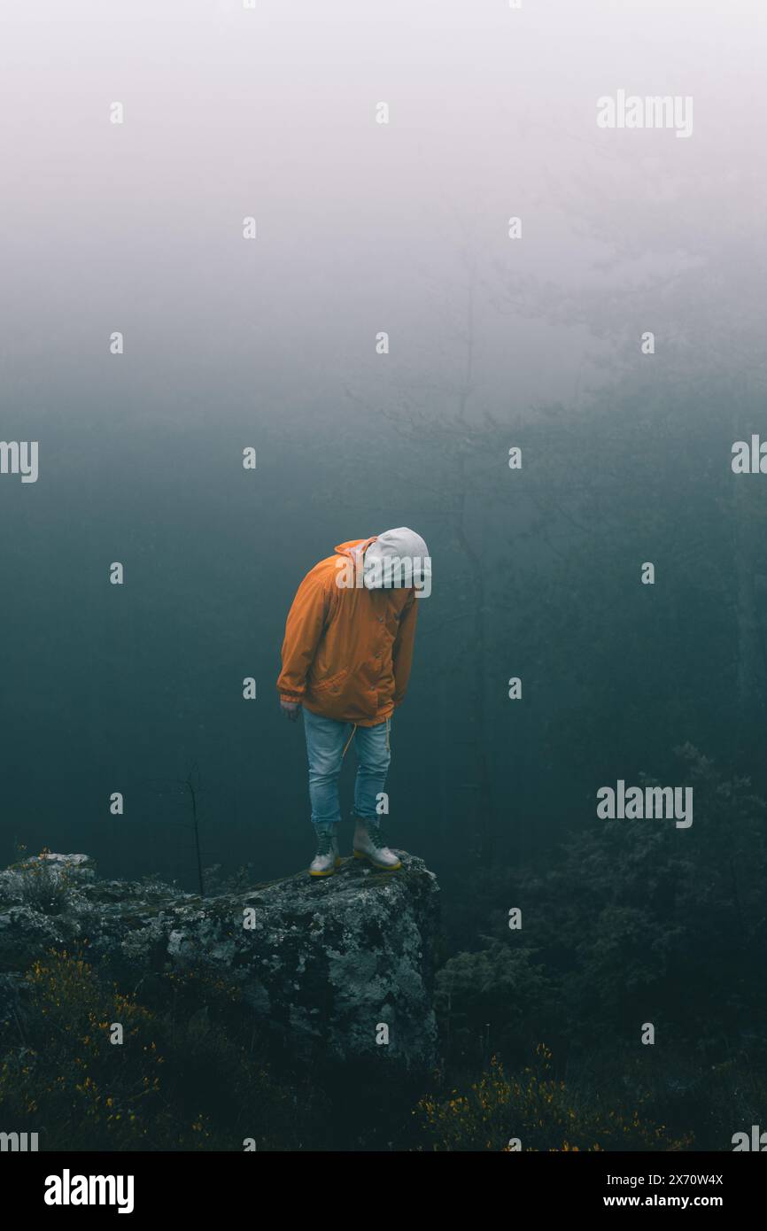 Bulgarien - 05 22 2024: Mann mit orangefarbener Jacke und blauer Jeans, der am Rand eines großen Felsens steht und die Klippe hinunter blickt mit dunklem, nebeligem und regnerischem B Stockfoto