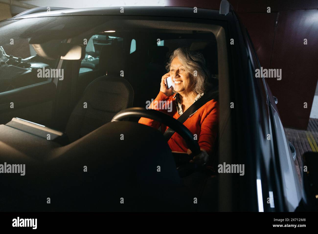 Eine ältere Frau, die in einem Auto sitzt, das in einer Tiefgarage geparkt ist und lächelt, während sie am Telefon spricht. Stockfoto