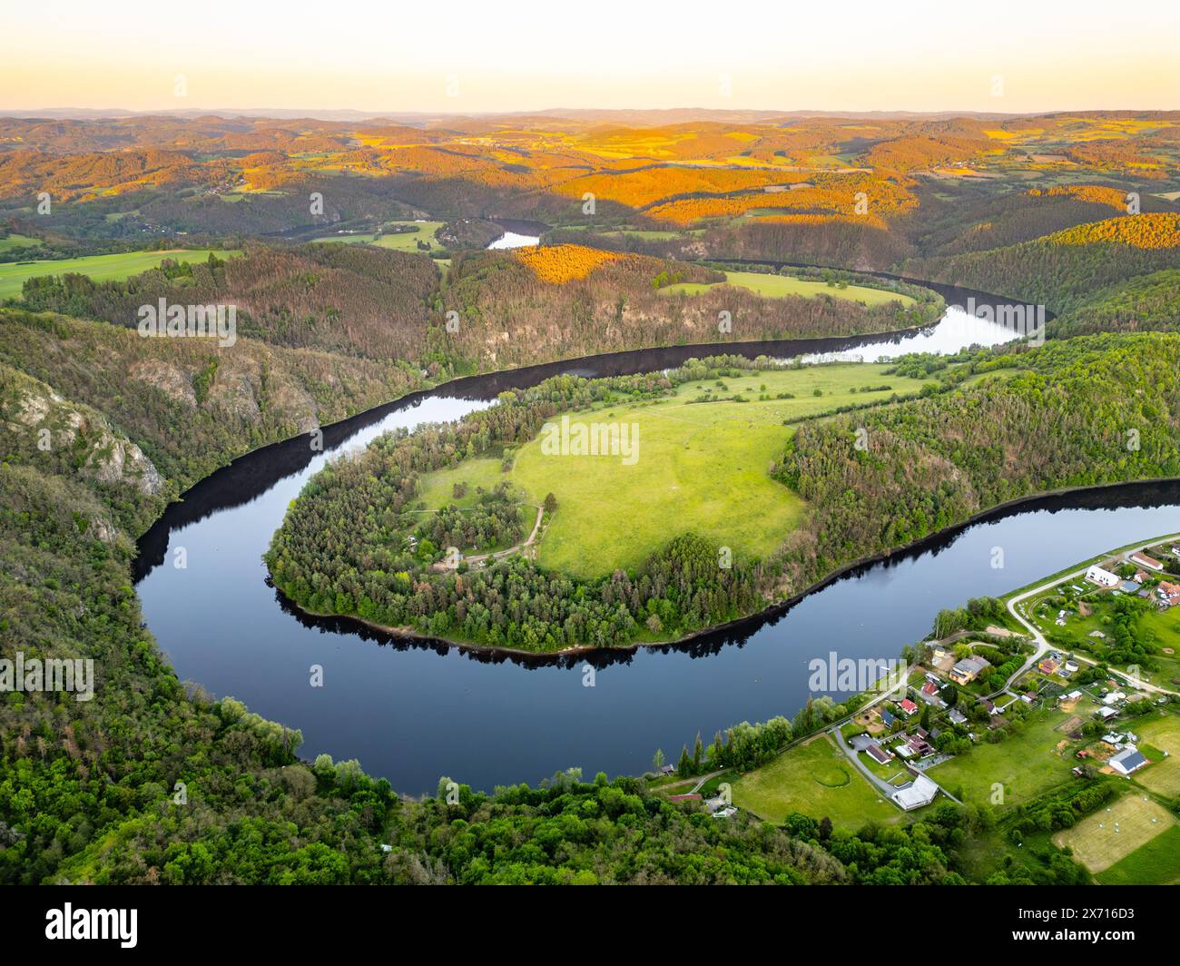 Blick aus der Vogelperspektive auf den Hufeisenbogen Solenice an der Moldau in Tschechien, mit Blick auf den sich windenden Fluss, umgeben von üppigem Grün und einem kleinen Dorf bei einem malerischen Sonnenuntergang. Stockfoto