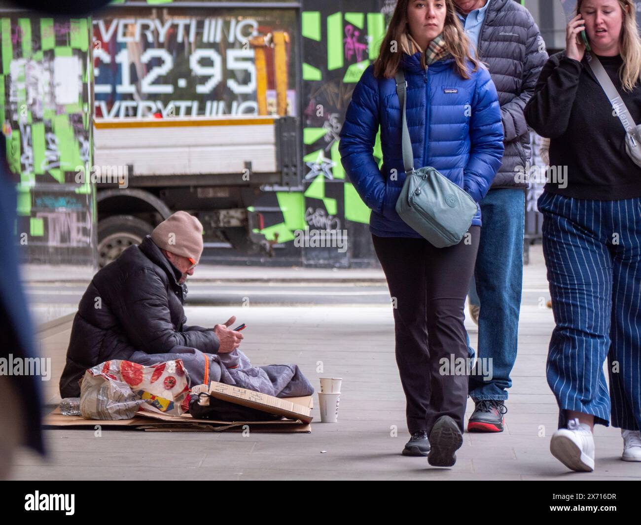 Obdachloser Mann im Schlafsack mit Handy unter den Menschenmassen von Einkäufern, Charing Cross Road London Stockfoto Obdachloser Mann im Schlafsack mit Handy unter den Menschenmassen von Einkäufern, Charing Cross Road London Stockfoto
