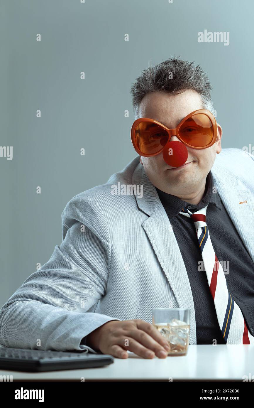 Ein Mann mittleren Alters in einem leichten Anzug und einer gestreiften Krawatte trägt eine übergroße orange Brille und eine rote Clownnnase, die ein Whiskyglas hält. Sein verspieltes, lächerliches Stockfoto