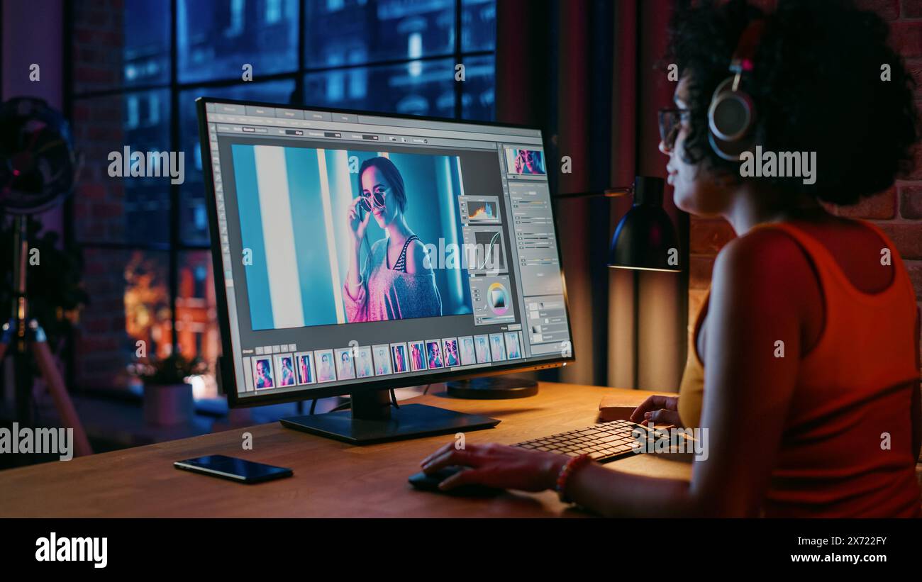 Junge kreative Frau in Kopfhörern mit Computer in stilvollem Loft Apartment am Abend. Grafikdesigner arbeitet von zu Hause aus, bearbeitet Modefotos für den Online-Shop. Blick auf die Stadt vom großen Fenster. Stockfoto