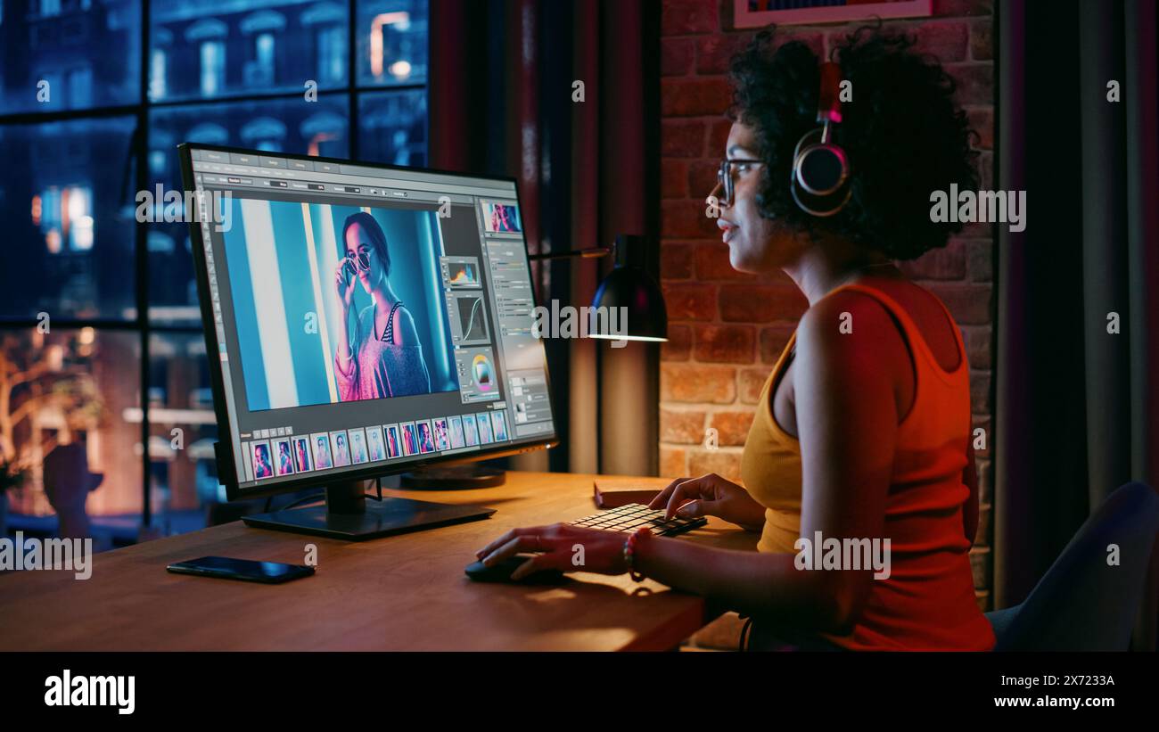 Junge kreative Frau in Kopfhörern mit Computer in stilvollem Loft Apartment am Abend. Grafikdesigner arbeitet von zu Hause aus, bearbeitet Modefotos für den Online-Shop. Blick auf die Stadt vom großen Fenster. Stockfoto