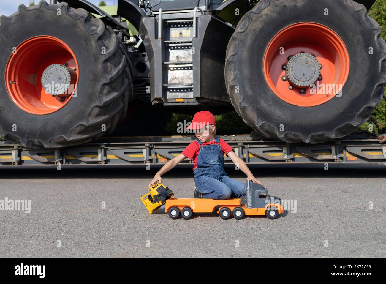 Junge spielt mit Spielzeugautos. Hinter ihm steht ein echter Traktor mit riesigen Rädern. Das Kind will Fahrer oder Mechaniker werden, wie Dad. Kommerzielle Fahrzeuge Stockfoto