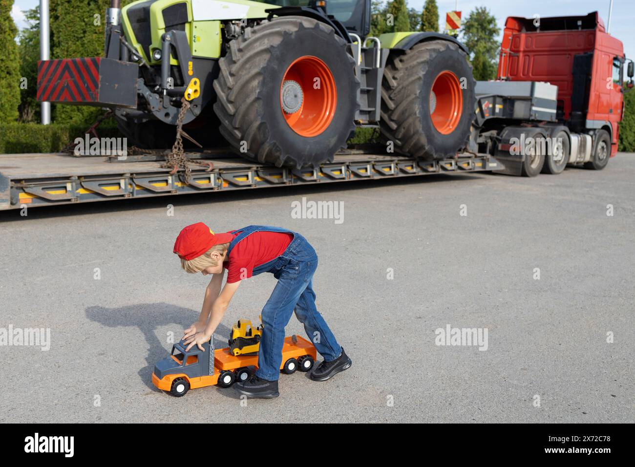 Junge spielt mit Spielzeugautos auf dem Parkplatz neben einem großen Truck mit beladenem Traktor. Das Kind träumt davon, Fahrer oder Mechaniker zu werden, wie Dad Stockfoto