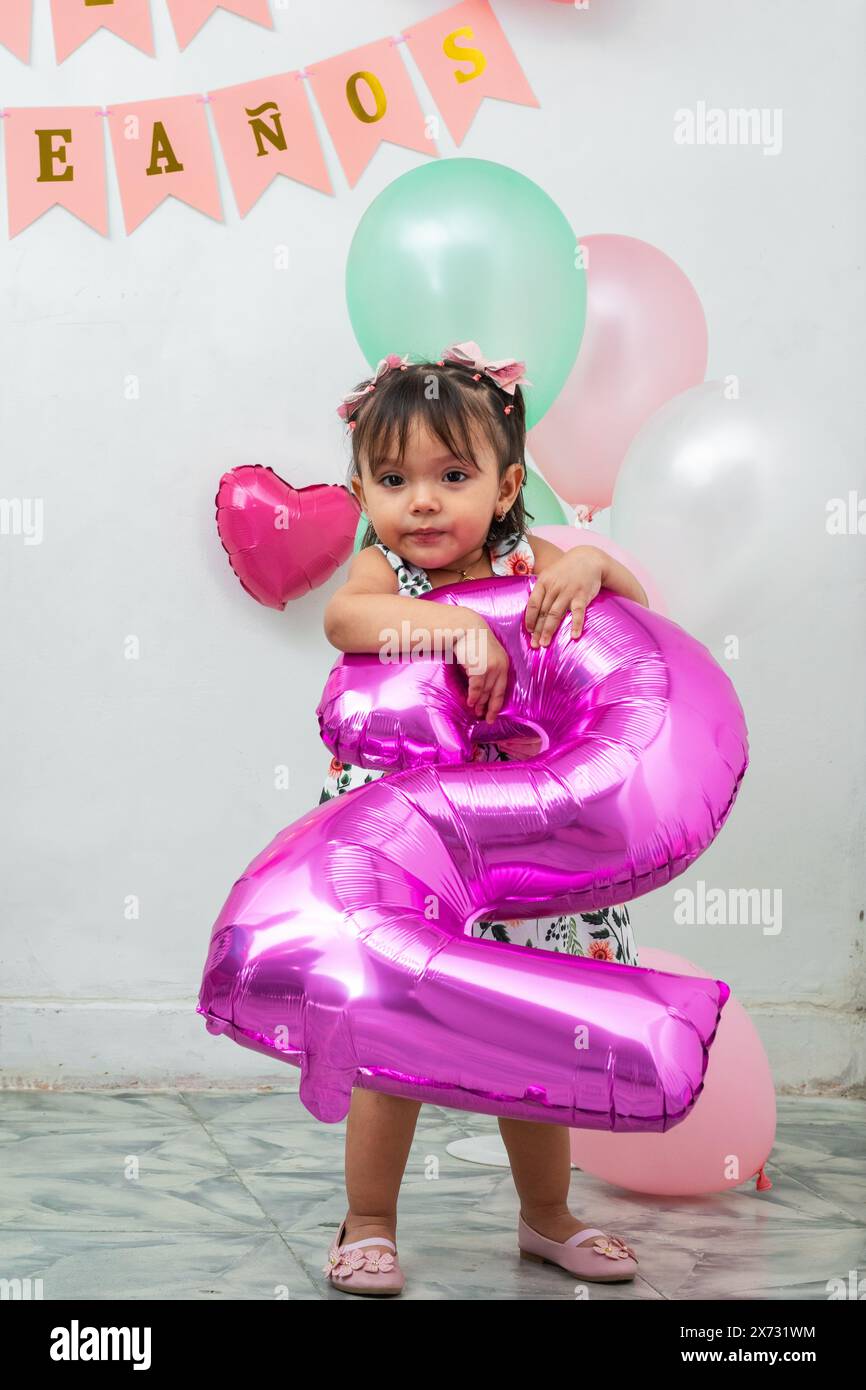 Das kleine brünette Latina-Mädchen hält einen fuchsiaförmigen Ballon mit Ballons hinter sich Stockfoto